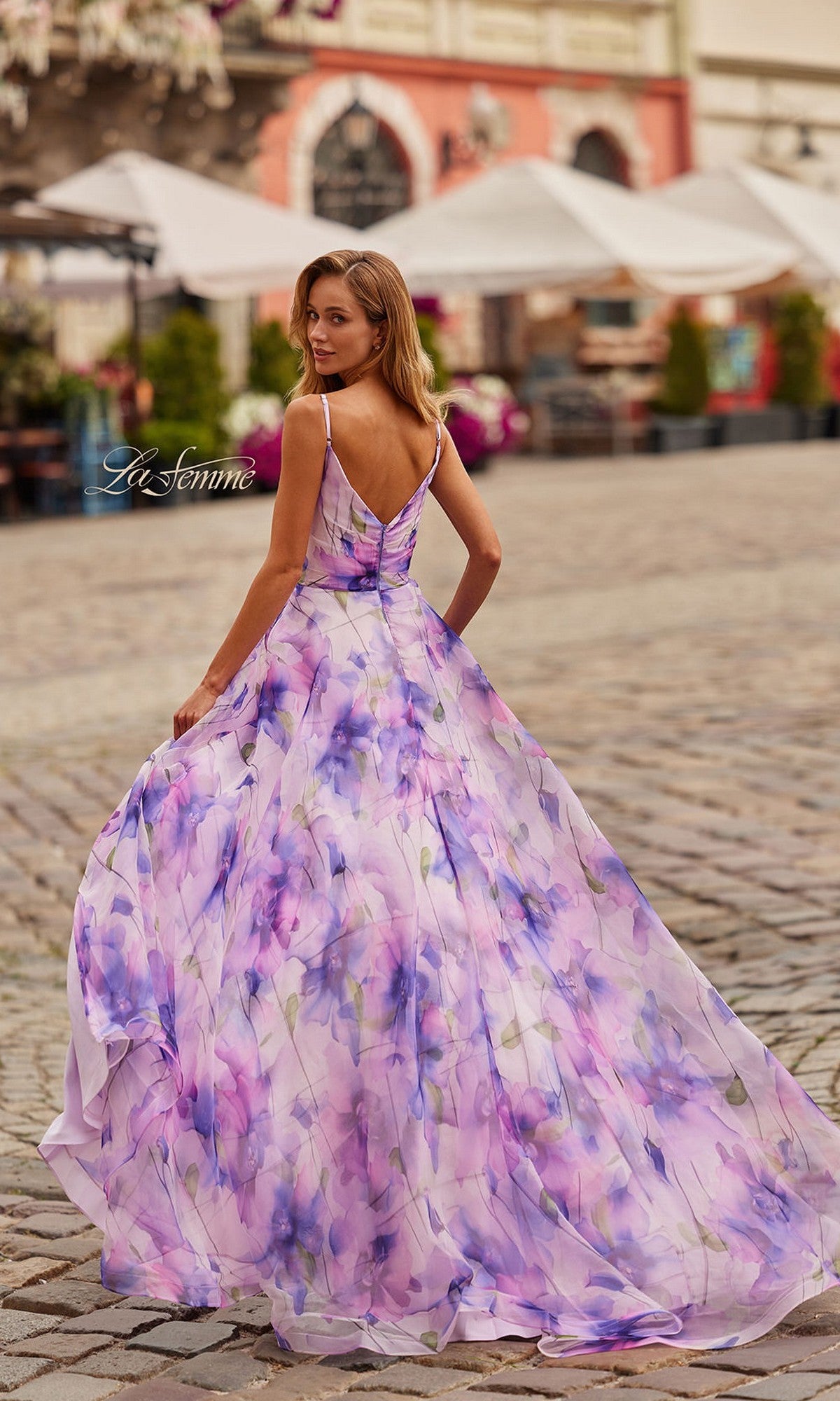 A woman wearing the La Femme 33606 long prom dress with a floral print and adjustable spaghetti straps stands on a cobblestone street, glancing over her shoulder amid outdoor cafés and historic buildings.